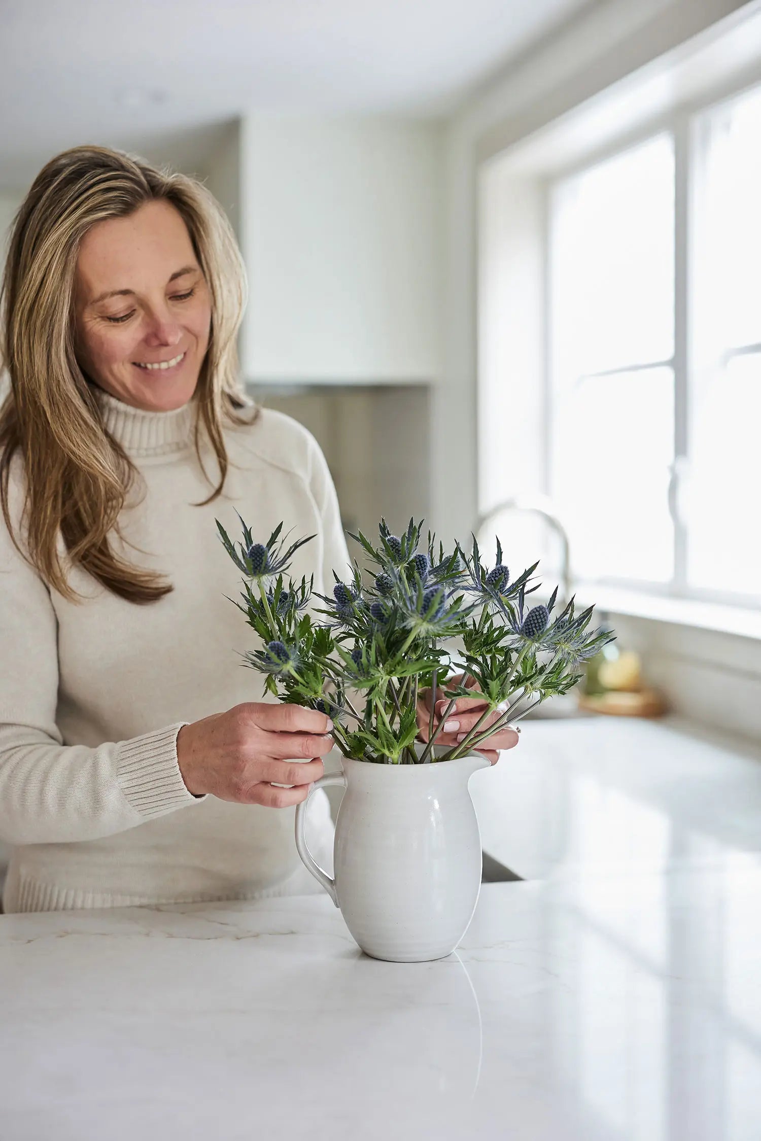Juniper Loft founder Christina arranges purple thistle in a white jug in a bright kitchen