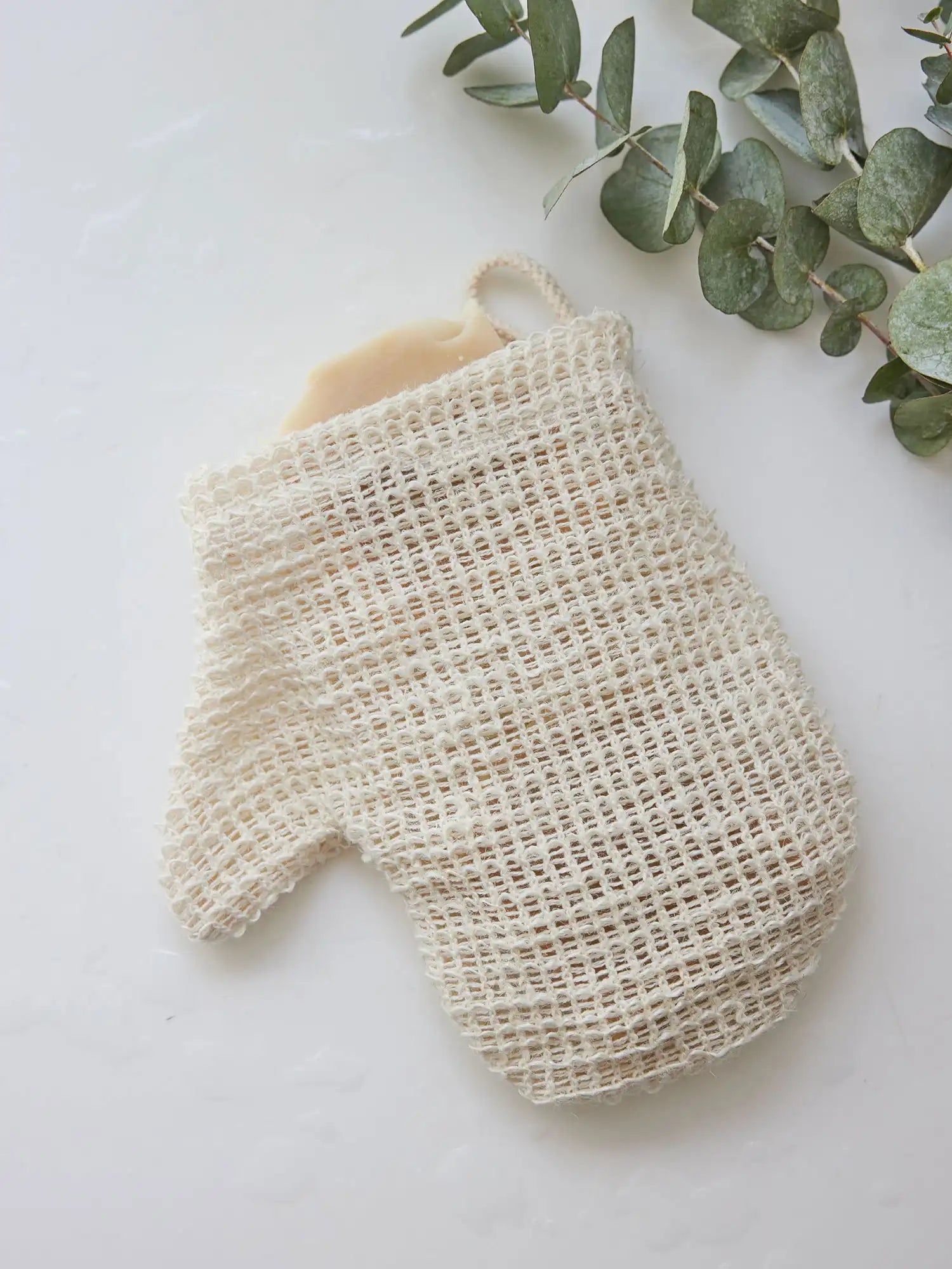 Sisal mitt from Juniper Loft Soap Company with a bar of soap inside next to eucalyptus leaves