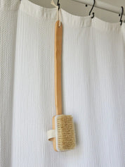 Long-handled bath brush from Juniper Loft Soap Company hanging from a white shower curtain