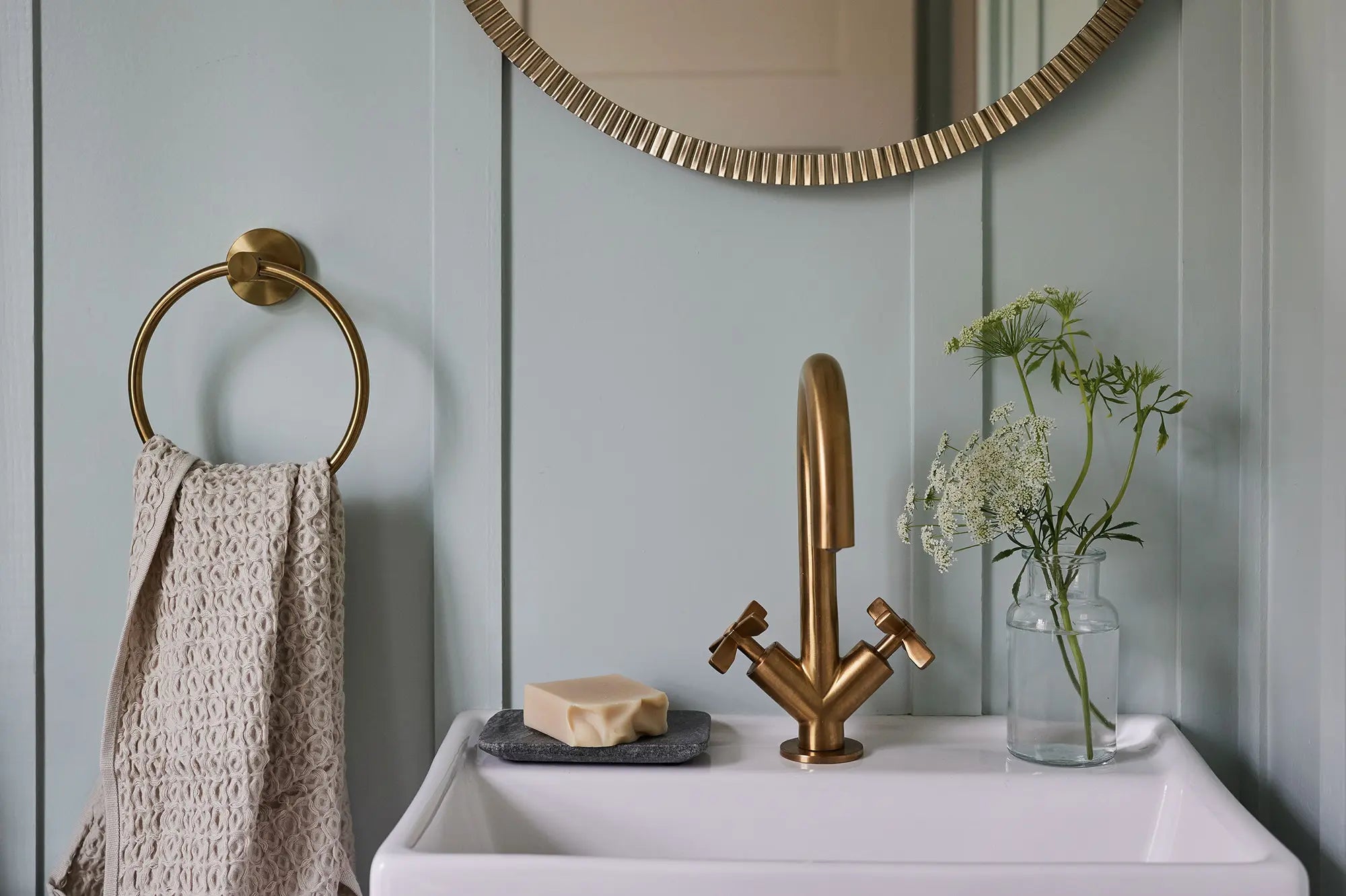 A gold bathroom faucet, mirror, towel, soap bar, and flowers on a white sink.