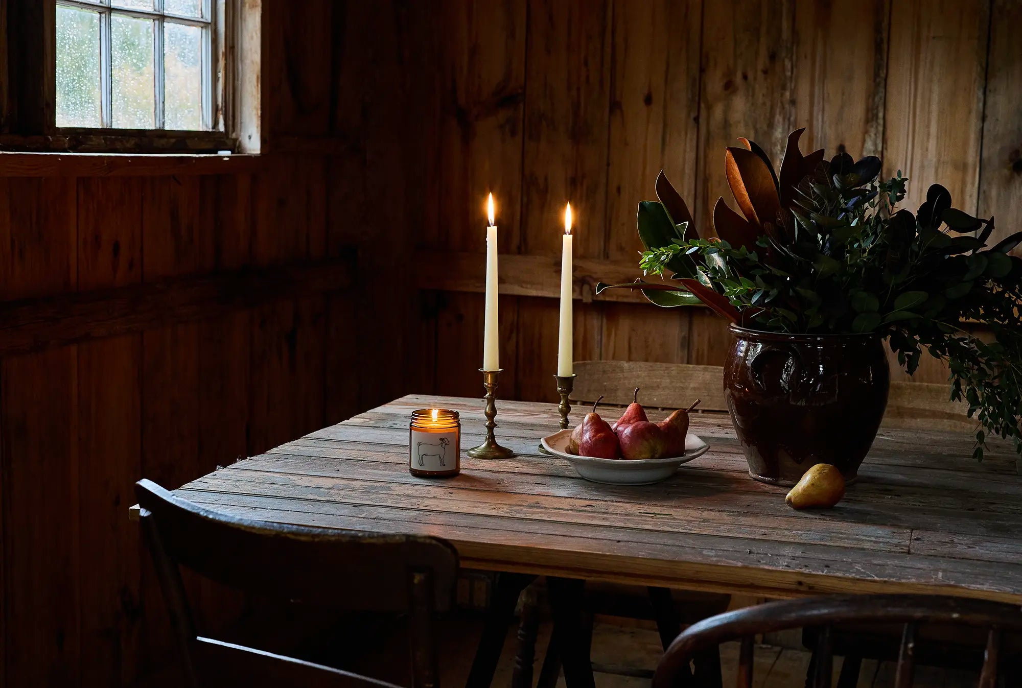 A large wooden table sits in a barn with candles, a bowl of red pears, and a vase with greenery
