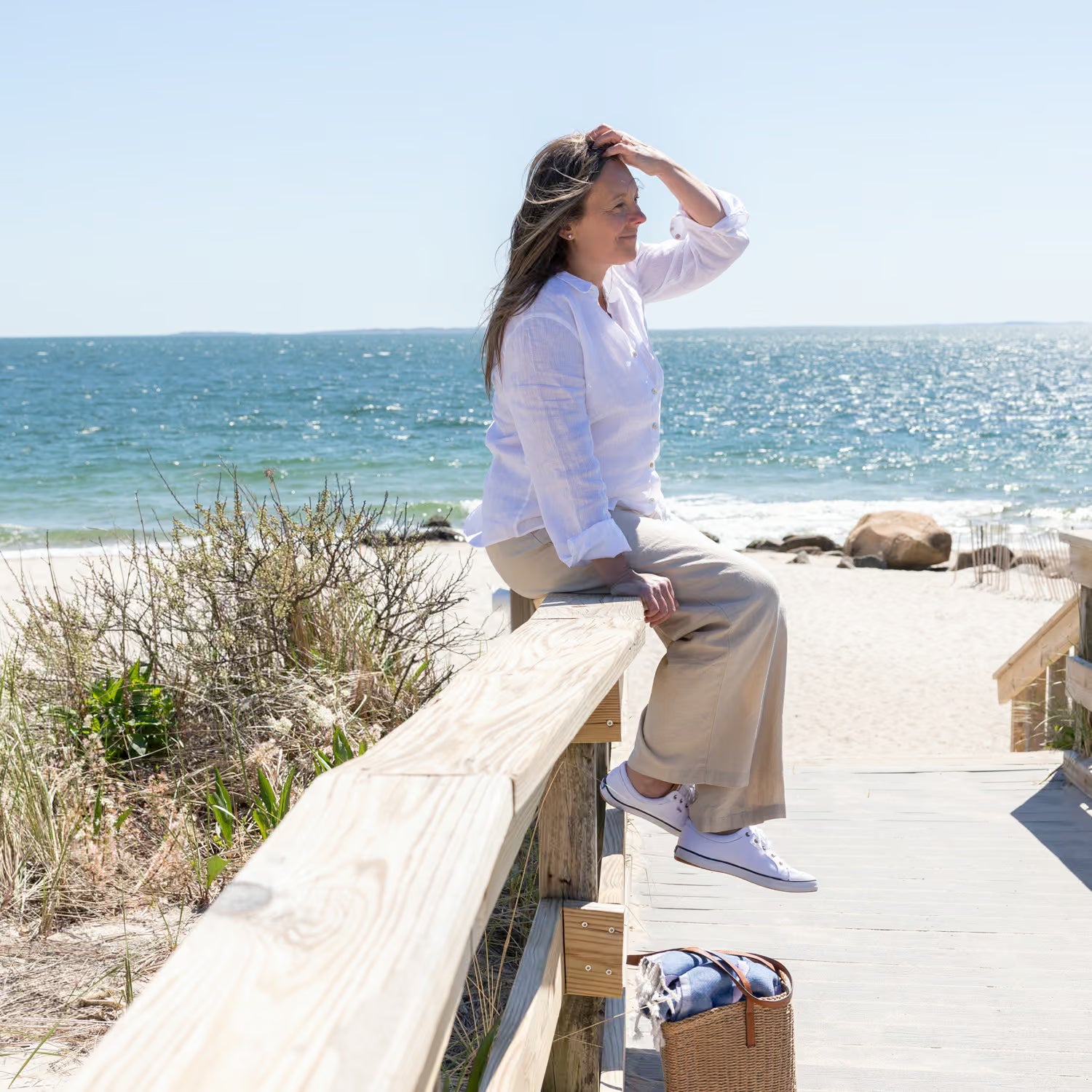 Juniper Loft founder Christina sitting on the wooden railing of a boardwalk at the  beach.