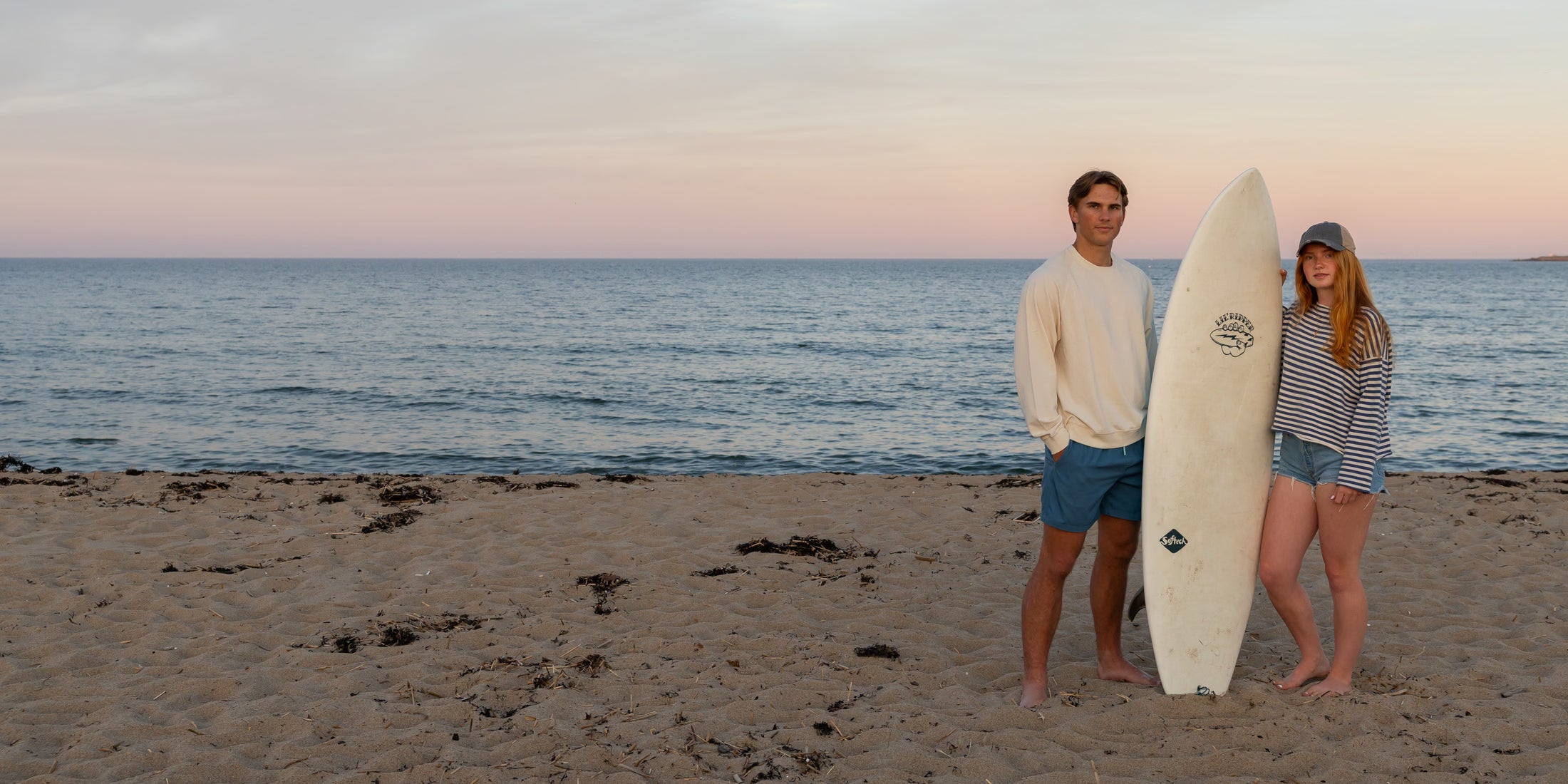 A man and a woman stand with a surfboard on a beach at sunset