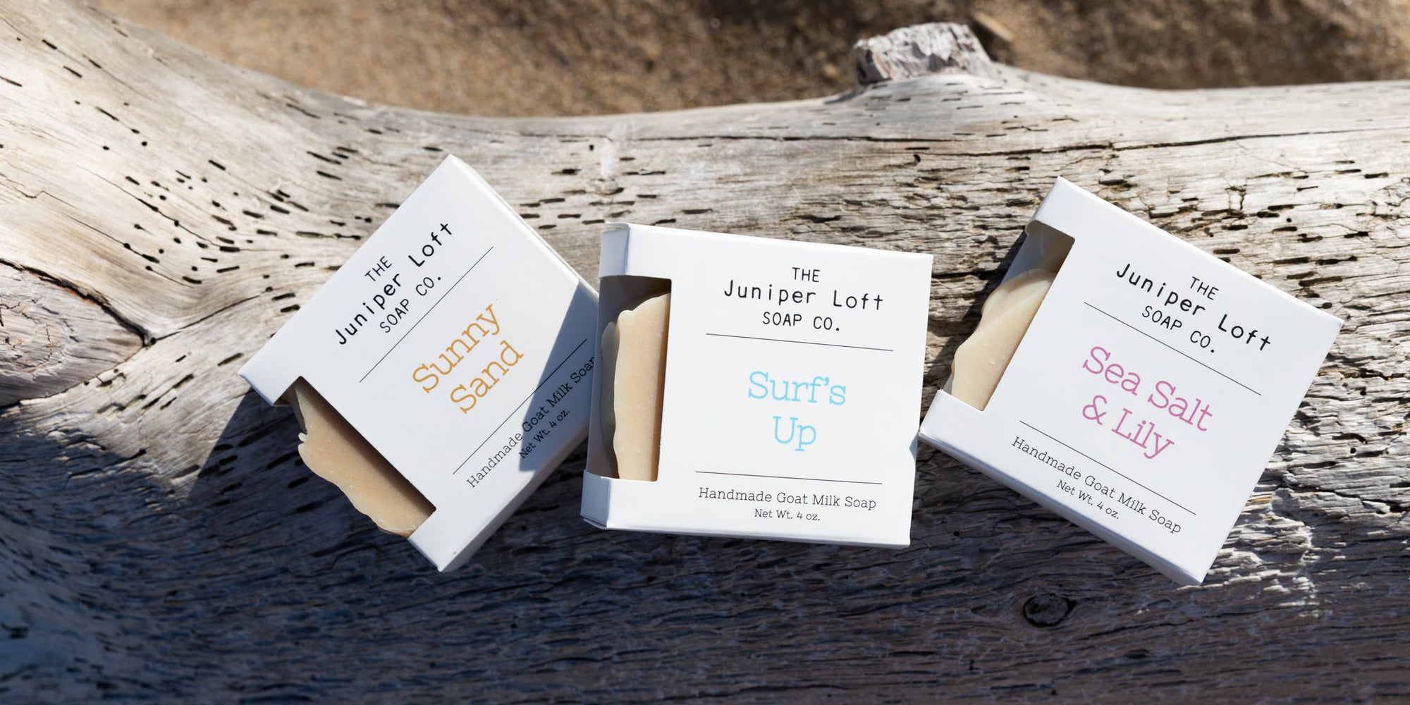 Three boxes of Juniper Loft Goat Milk Soap rest on a large piece of driftwood