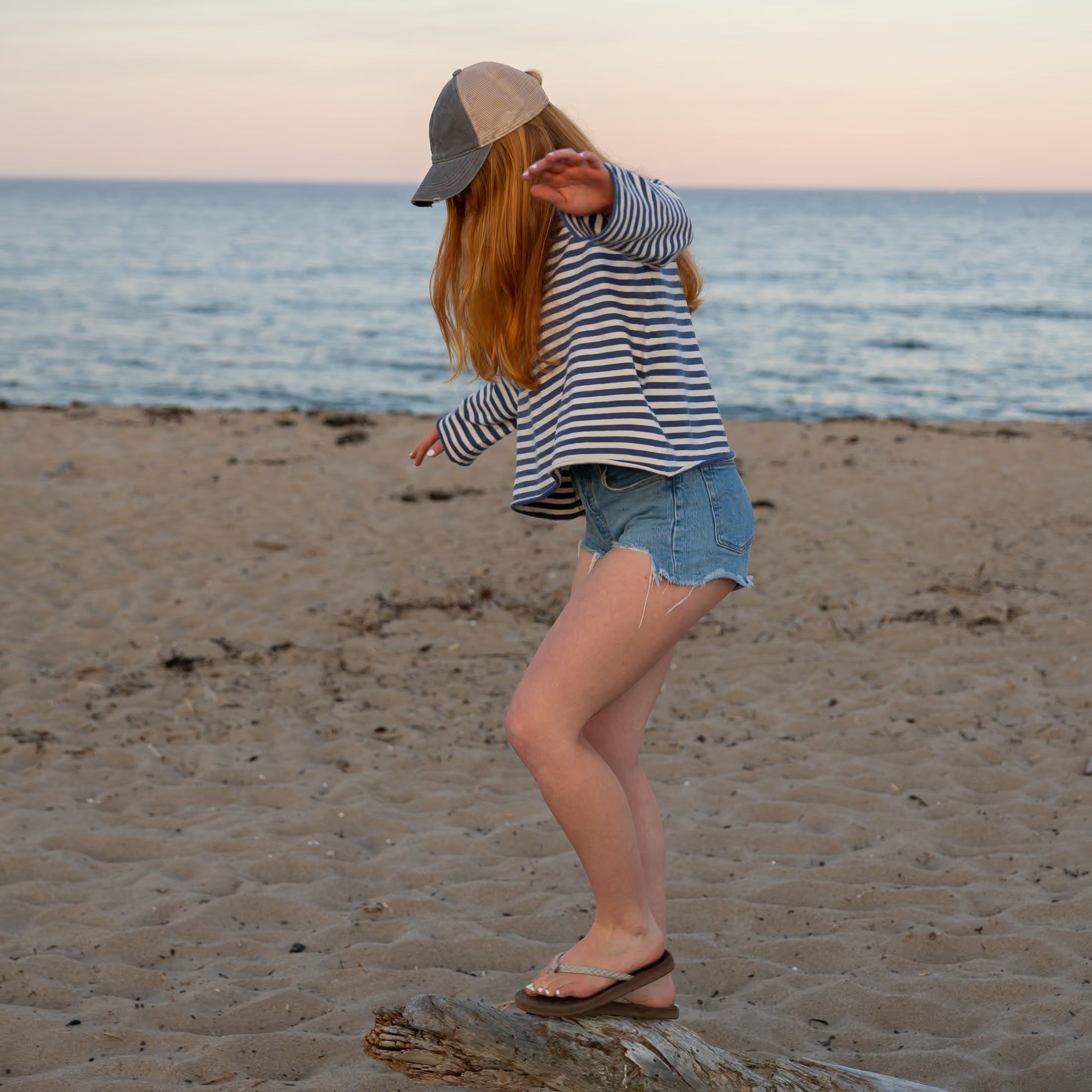 A woman wearing a cap, striped long-sleeve shirt and denim shorts balances on driftwood at the beach