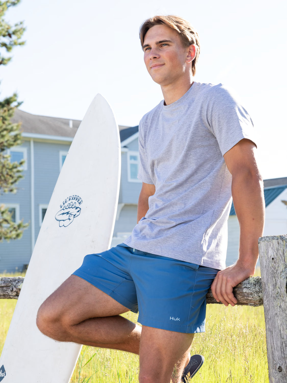 A man wearing a gray shirt and blue shorts leans against a wooden fence with his surfboard