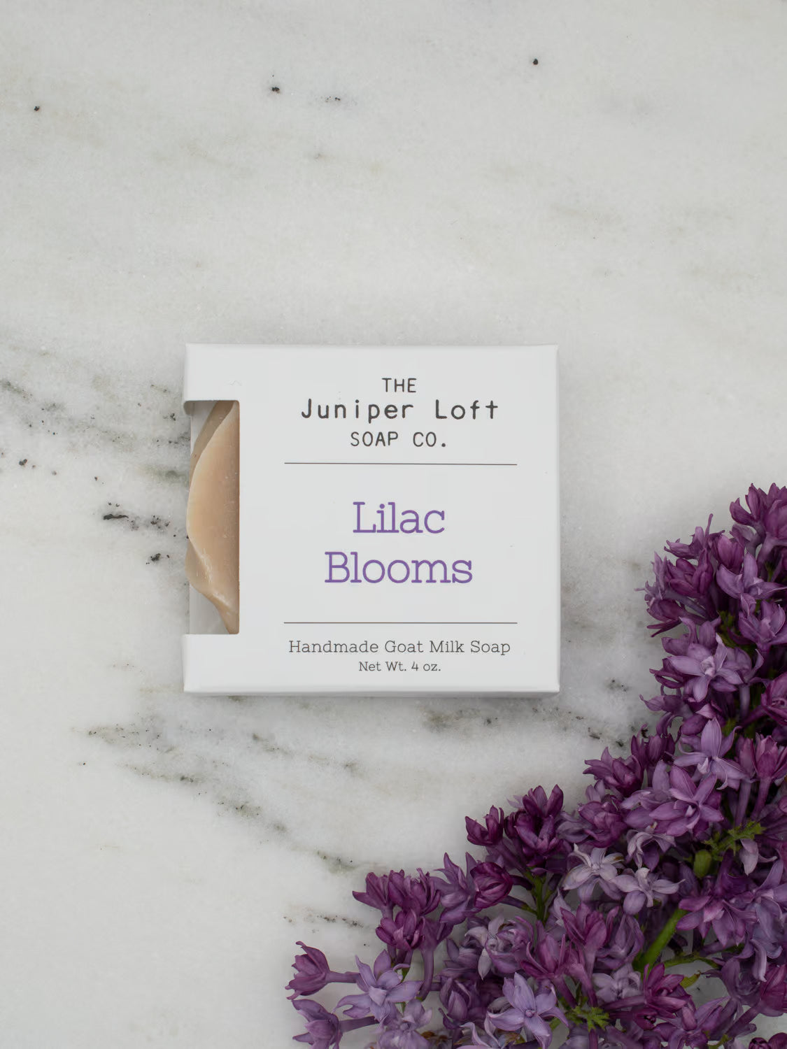 A bar of Juniper Loft Lilac Blooms soap on a marble surface with a sprig of purple Lilacs.