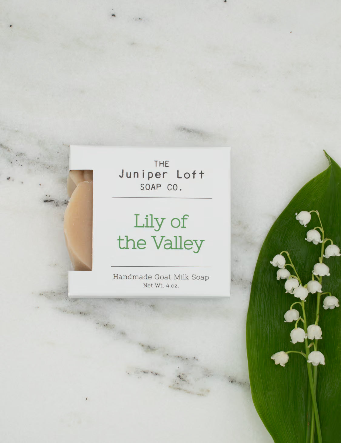 A bar of Lily of the Valley scented Juniper Loft goat milk soap set on a marble surface with a sprig of Lily of the Valley flowers.