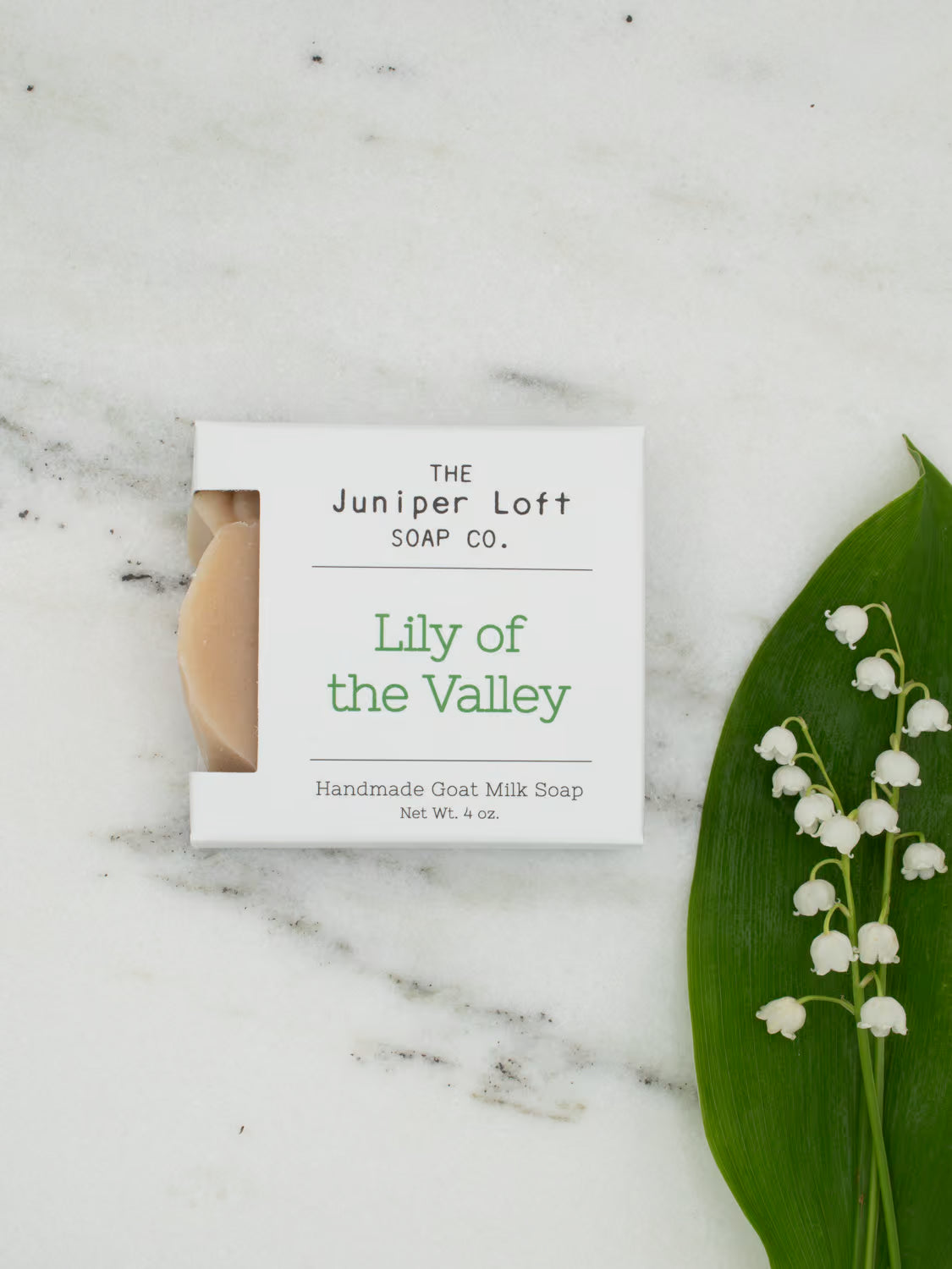 A bar of Lily of the Valley scented Juniper Loft goat milk soap set on a marble surface with a sprig of Lily of the Valley flowers.