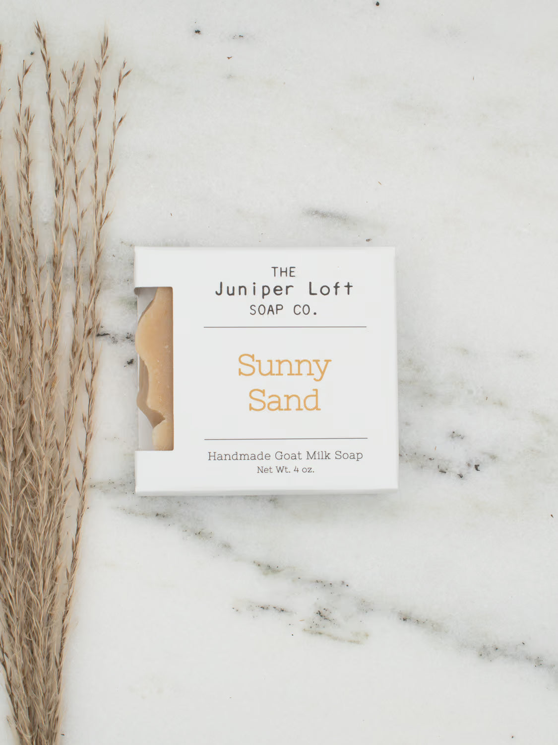 A square white soap bar package labeled 'THE Juniper Loft SOAP CO.' and 'Sunny Sand' sits on a white marble surface next to dried grass stems.
