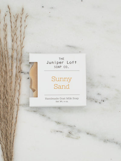 A square white soap bar package labeled 'THE Juniper Loft SOAP CO.' and 'Sunny Sand' sits on a white marble surface next to dried grass stems.