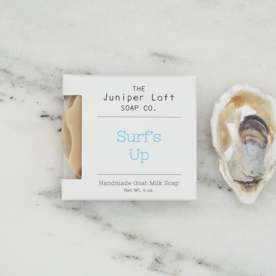 A square bar of The Juniper Loft Soap Company soap with the scent name Surf's Up on a marble slab with a shell alongside it.