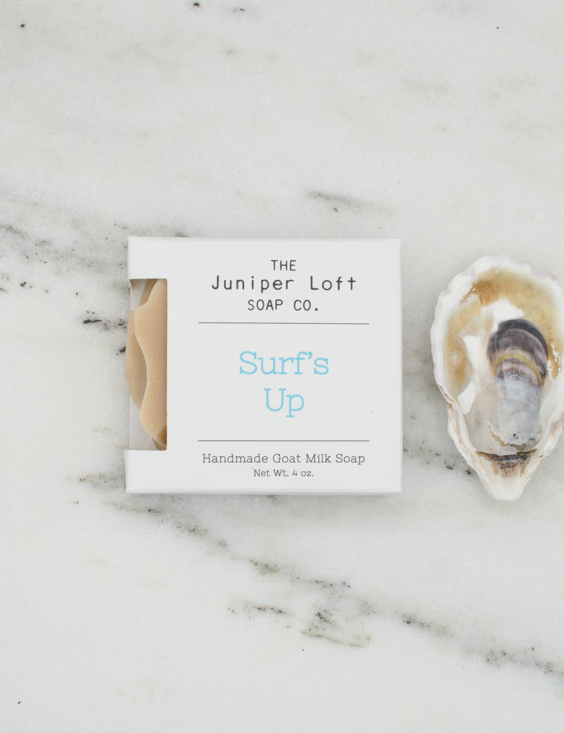 A square bar of The Juniper Loft Soap Company soap with the scent name Surf's Up on a marble slab with a shell alongside it.