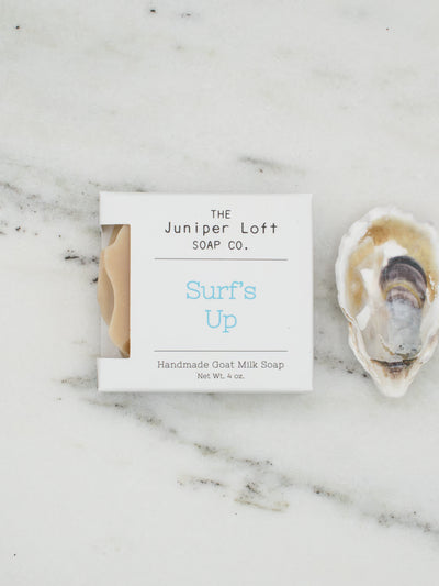A square bar of The Juniper Loft Soap Company soap with the scent name Surf's Up on a marble slab with a shell alongside it.