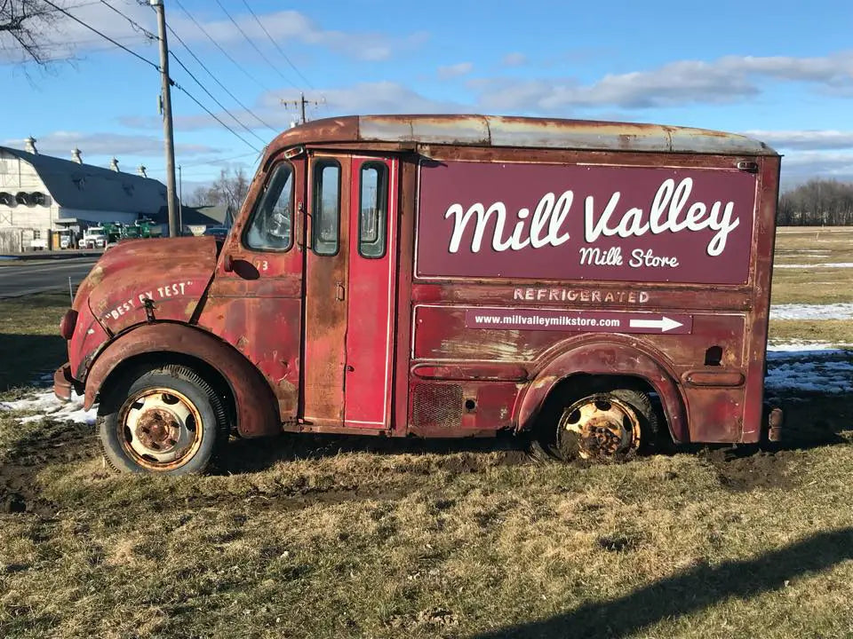 Rustic red truck with &#39;Mill Valley Milk Store&#39; branding on a grassy field.