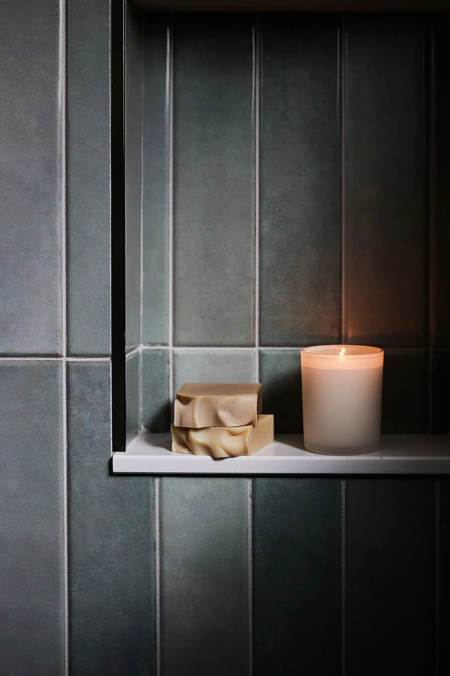 Candle and two bars of soap on a shower ledge with dark green tile