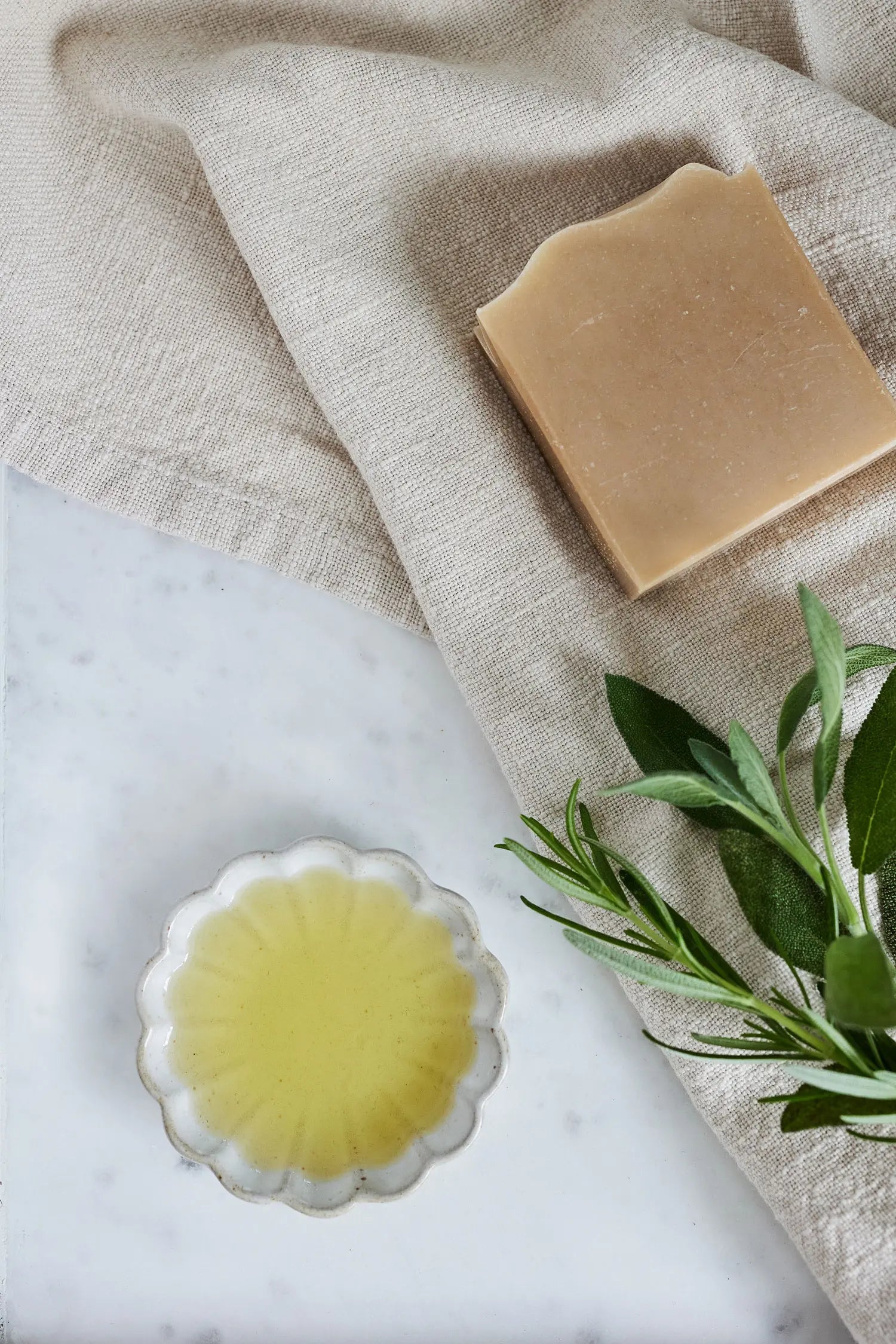 Bar of soap and a flower dish with essential oil on a marble surface with a beige towel and green leaves